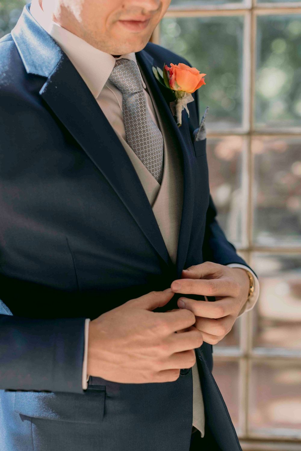 Close-up of groom in navy suit with gray tie and orange rose boutonniere fastening his jacket button.