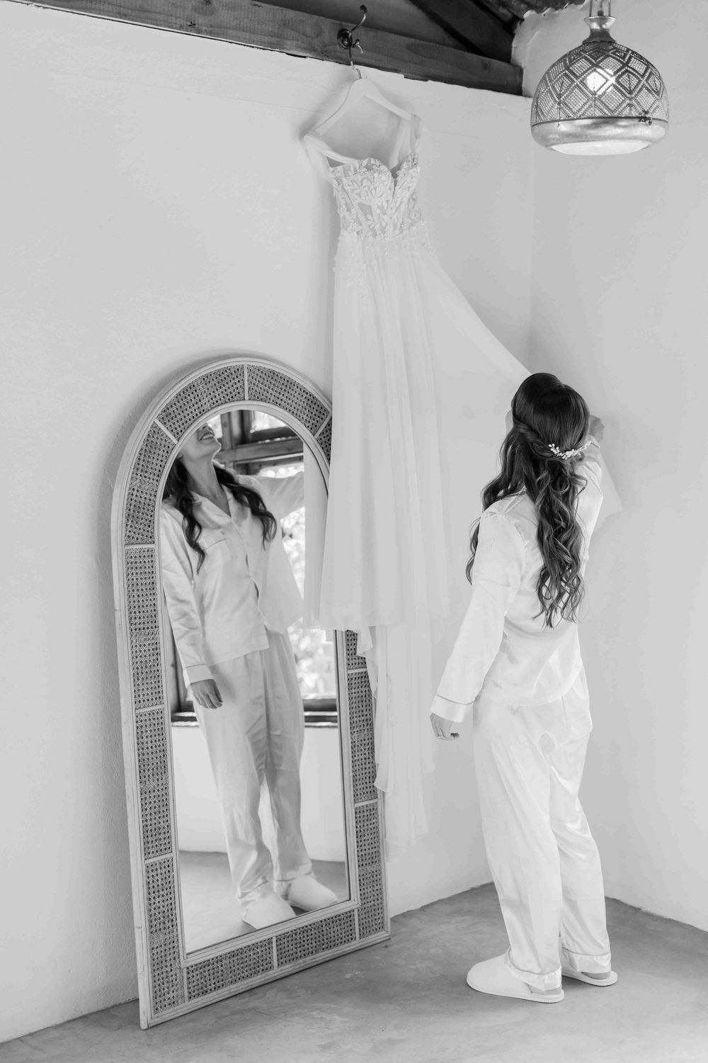 Bride in white pajamas standing before arched rattan mirror admiring hanging wedding dress, black and white photo.