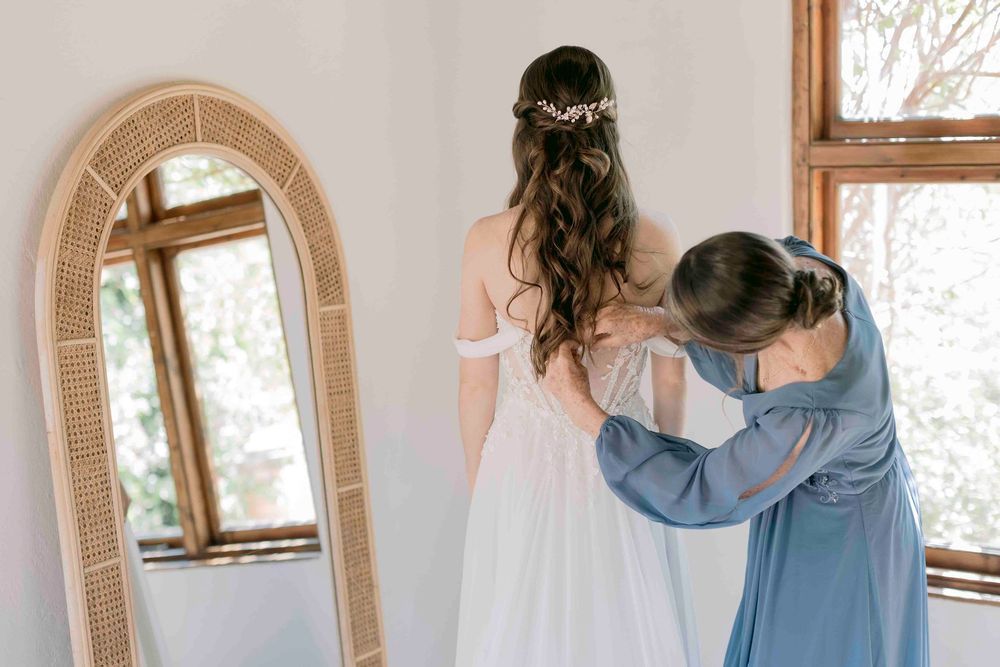 Helper fastening the back of bride's white lace off-shoulder wedding gown near arched mirror.