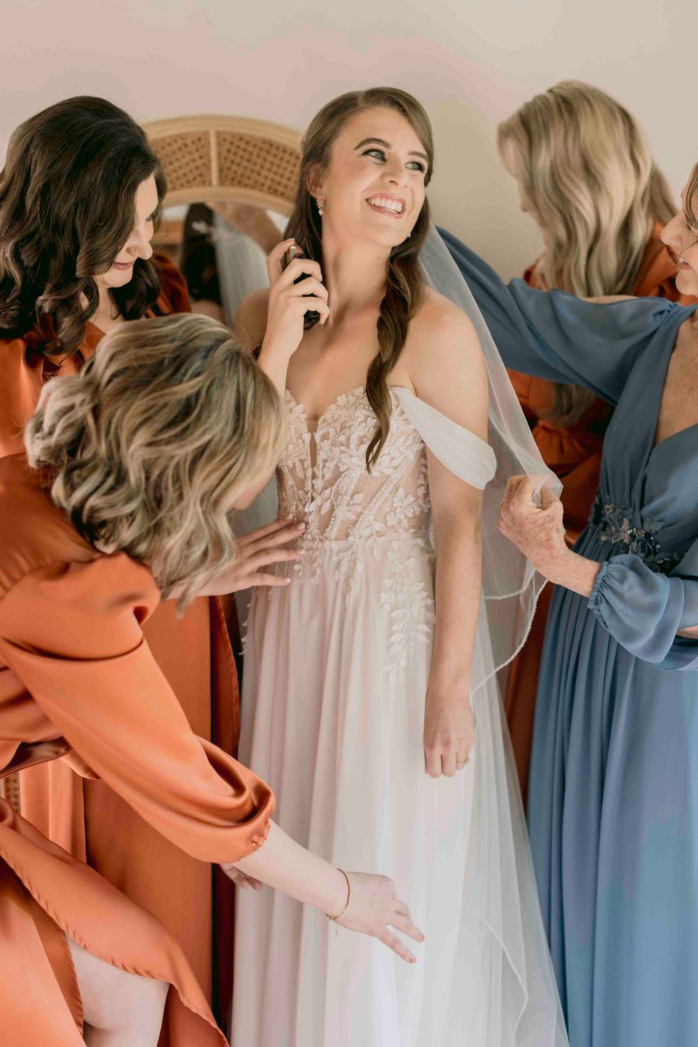 Smiling bride in lace gown surrounded by bridesmaids in rust and blue dresses.