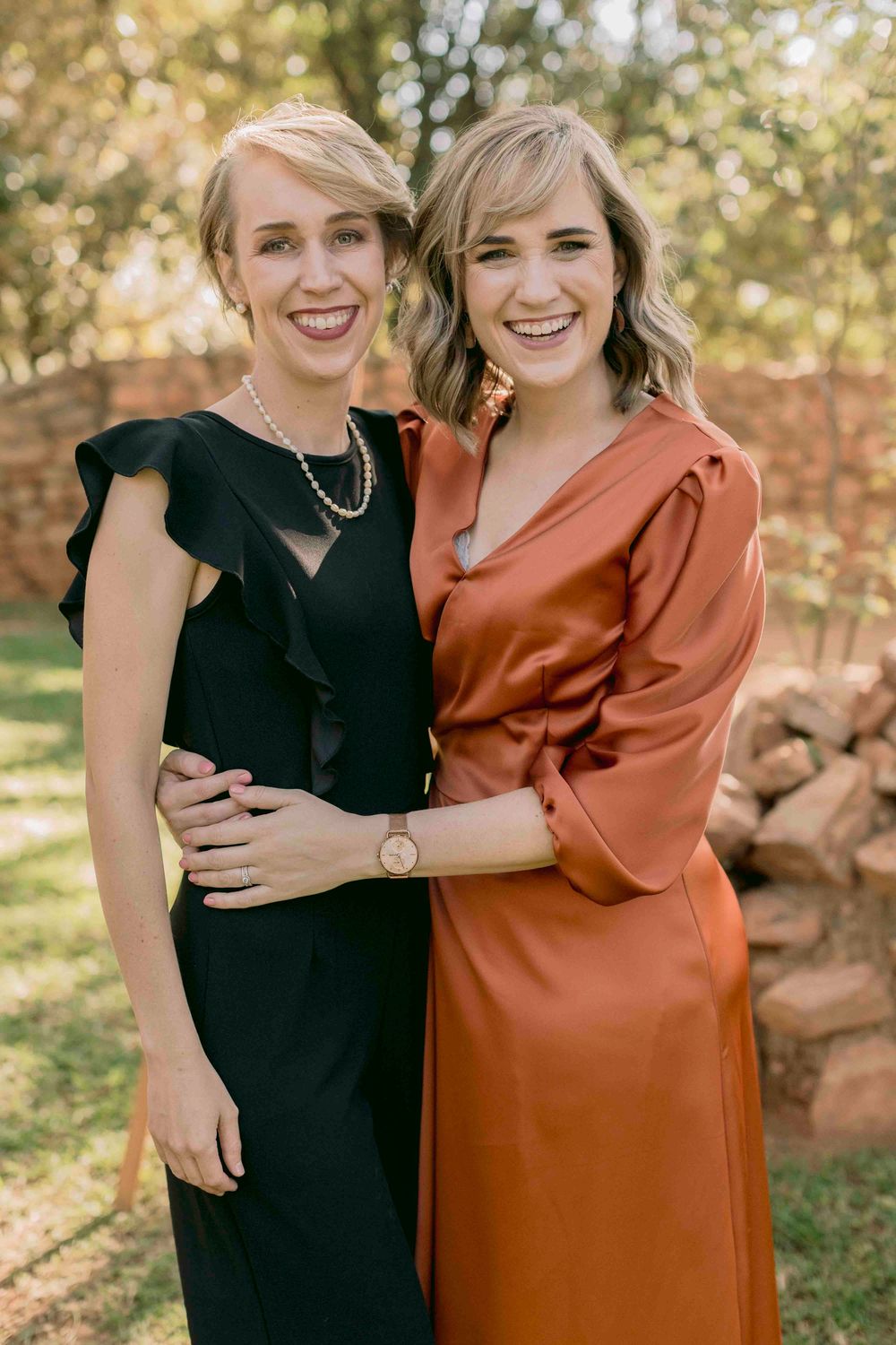 Two women smiling and posing together at an outdoor event, one in black and one in rust satin dress.