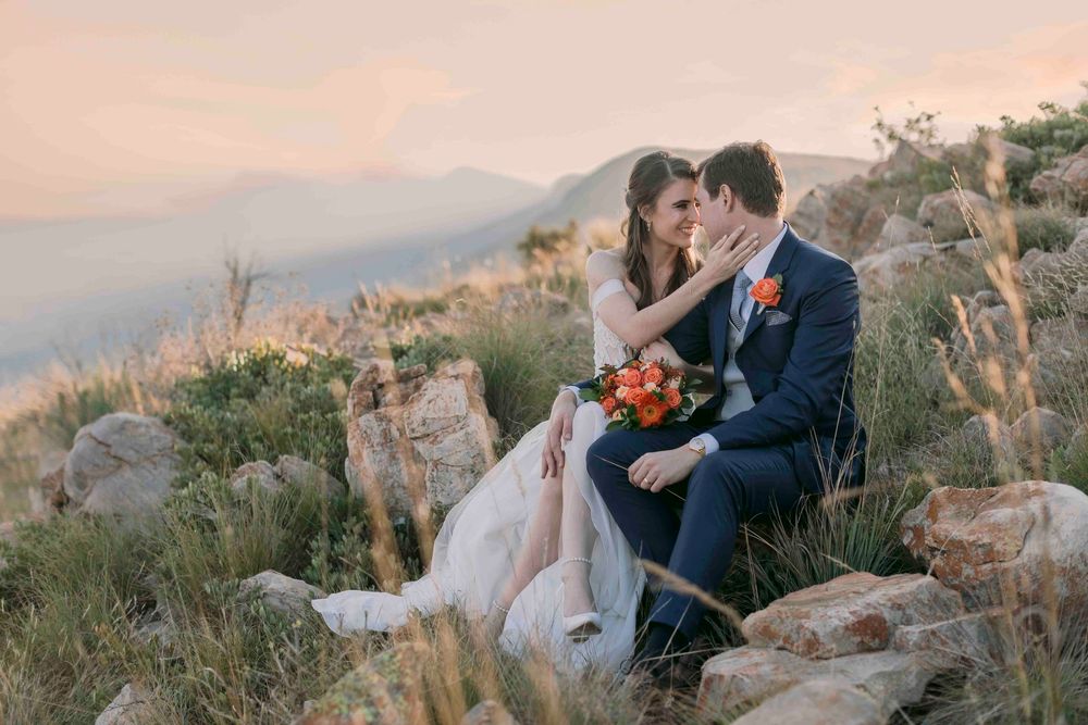Couple in wedding attire share an intimate moment seated on rocks with mountain landscape at golden hour.