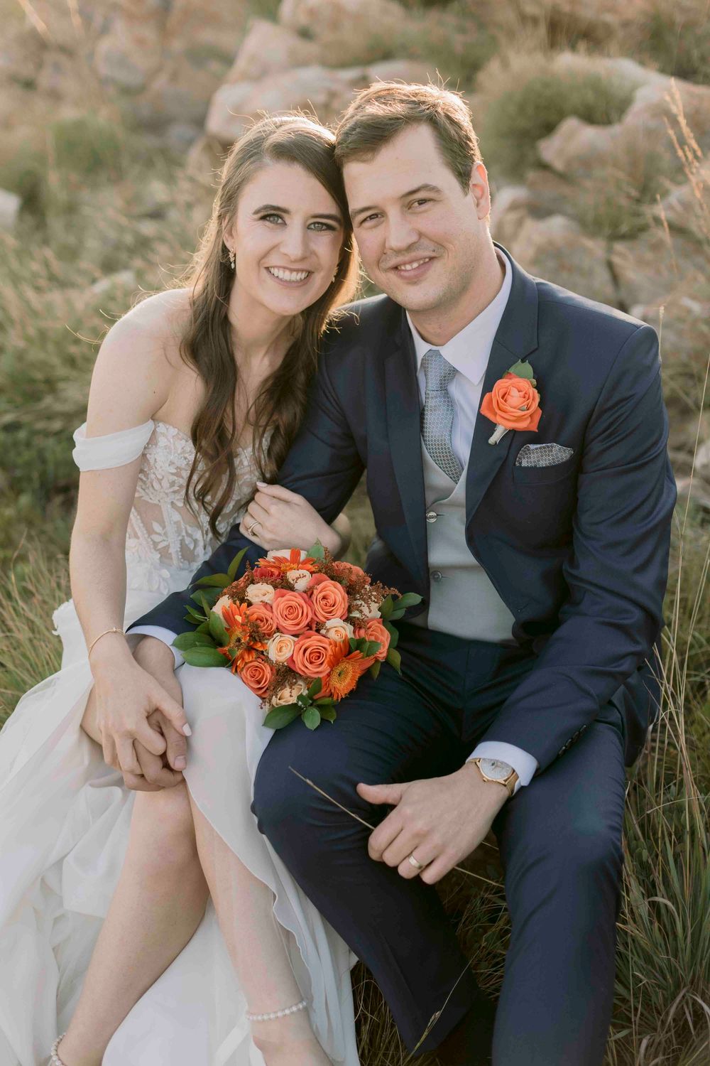 Happy bride and groom pose together on rocks, bride holding orange rose bouquet with mountain scenery behind them.