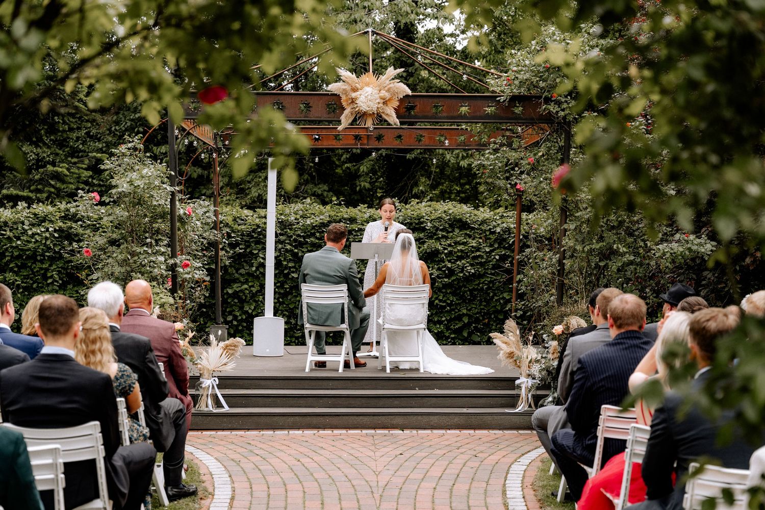 A scenic outdoor wedding ceremony setup with rows of chairs on a curved brick pathway surrounded by lush greenery.