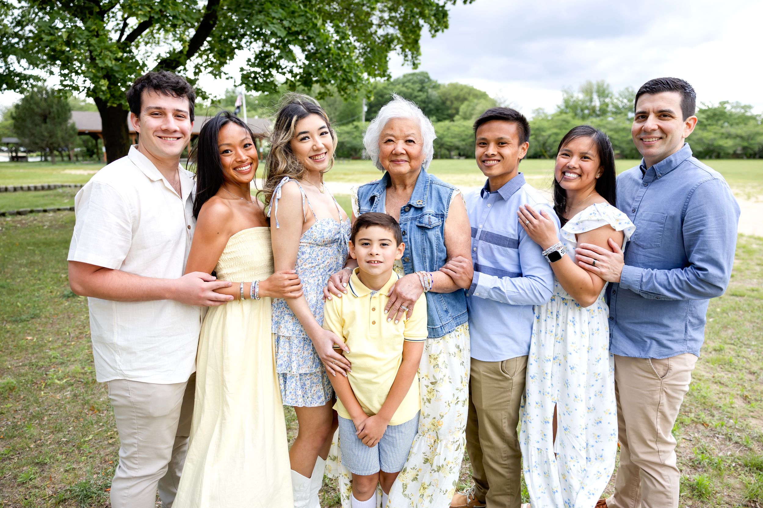 A multi-generational family smiles together outdoors on a sunny day in casual spring attire against a green background. Family NJ photograph