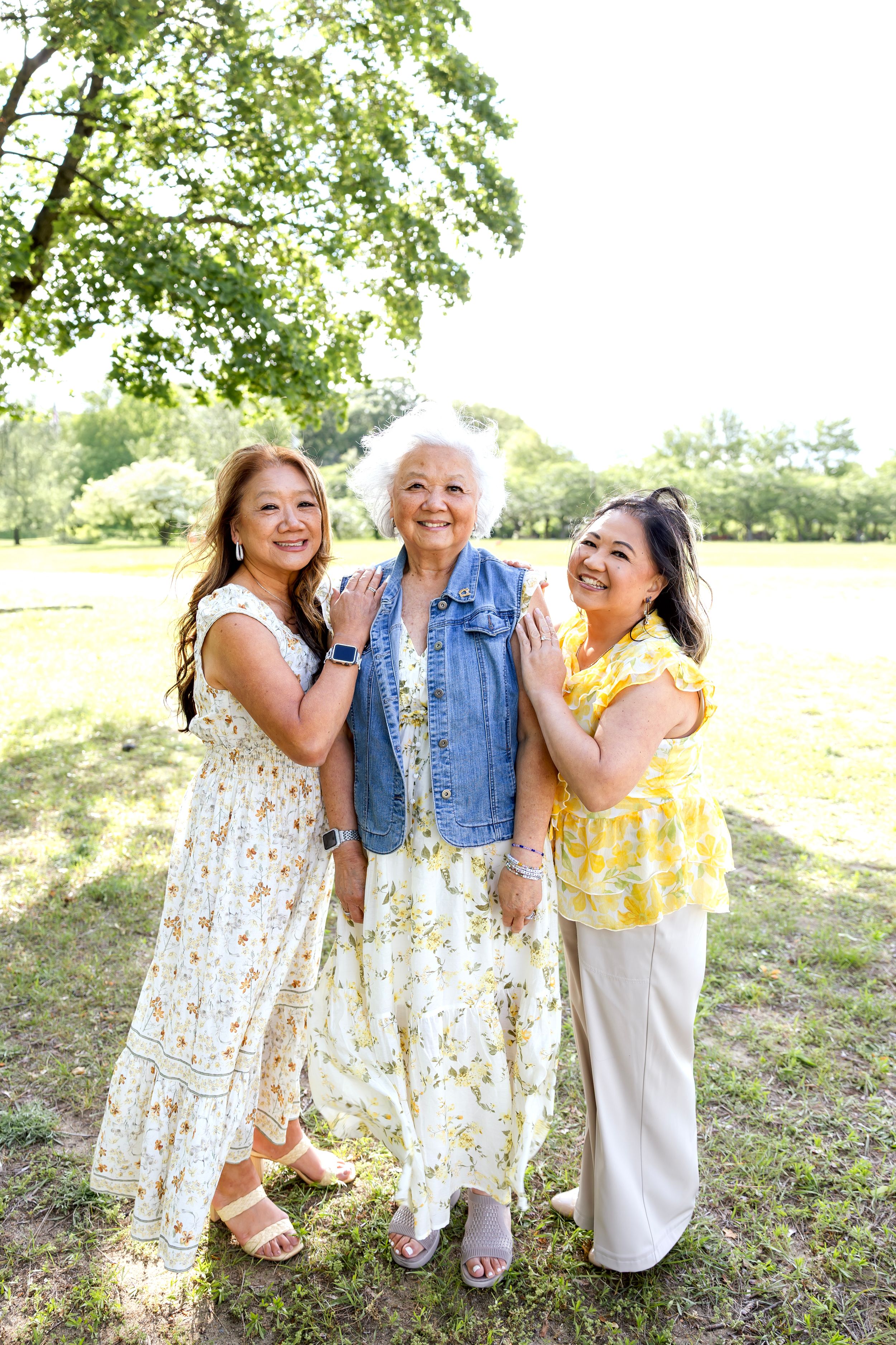 Three women embrace and laugh joyfully together on a grassy hillside in summer sunlight. Family NJ photographer