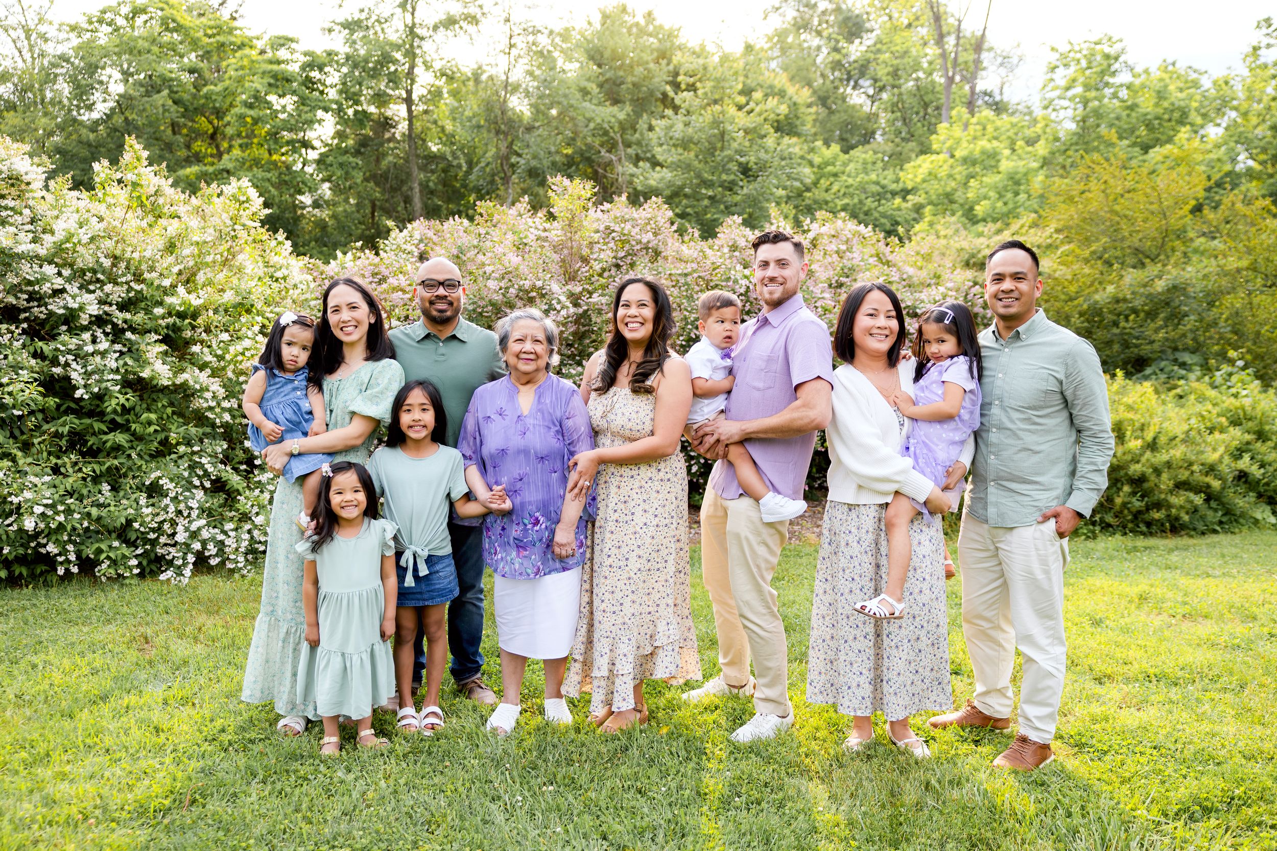 Extended family wearing coordinated pastel outfits poses together on grassy hill during outdoor photo session. Family NJ photographer