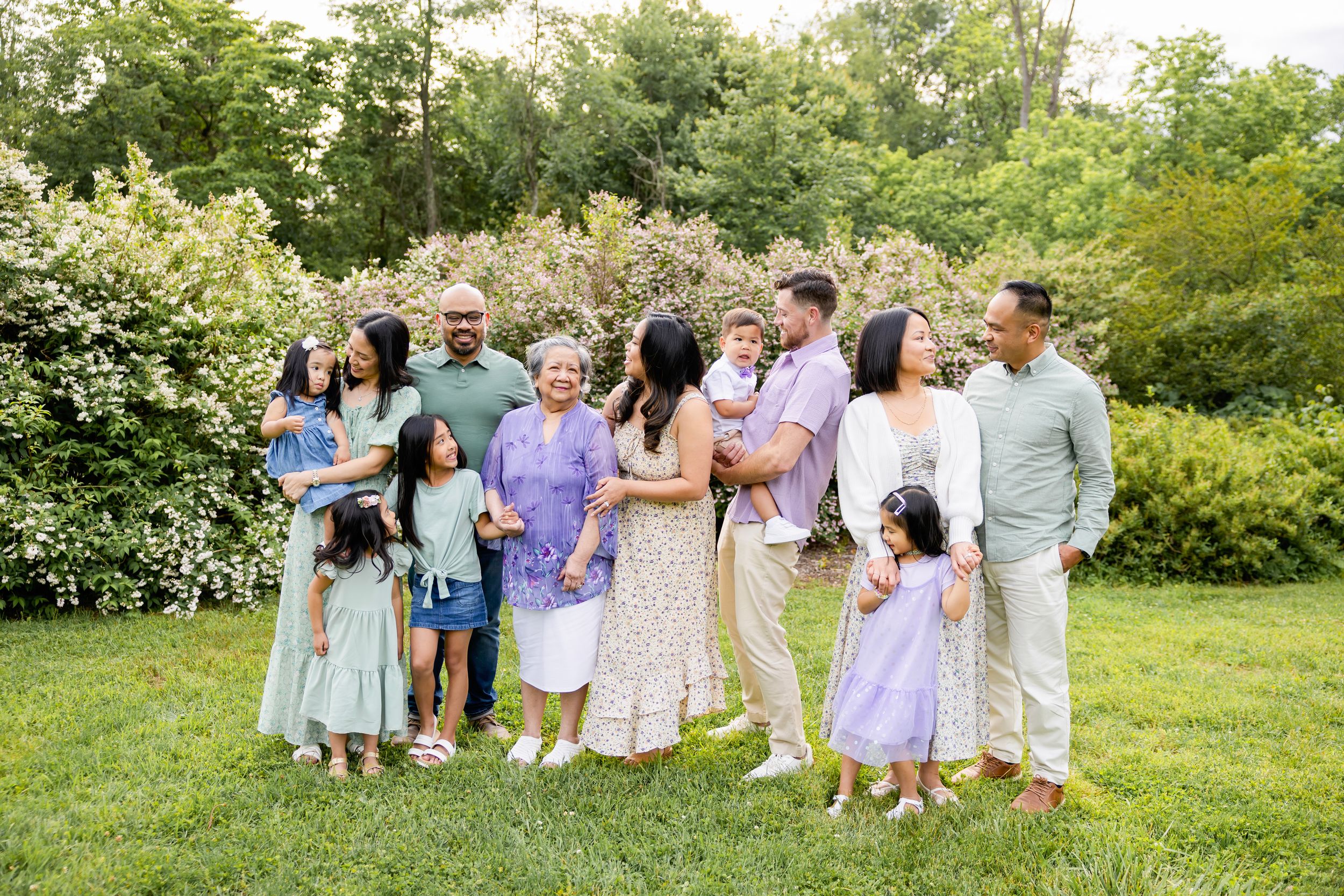 A group of people in coordinated pastel outfits sitting and standing together on green grass for outdoor photos. Family NJ photographer