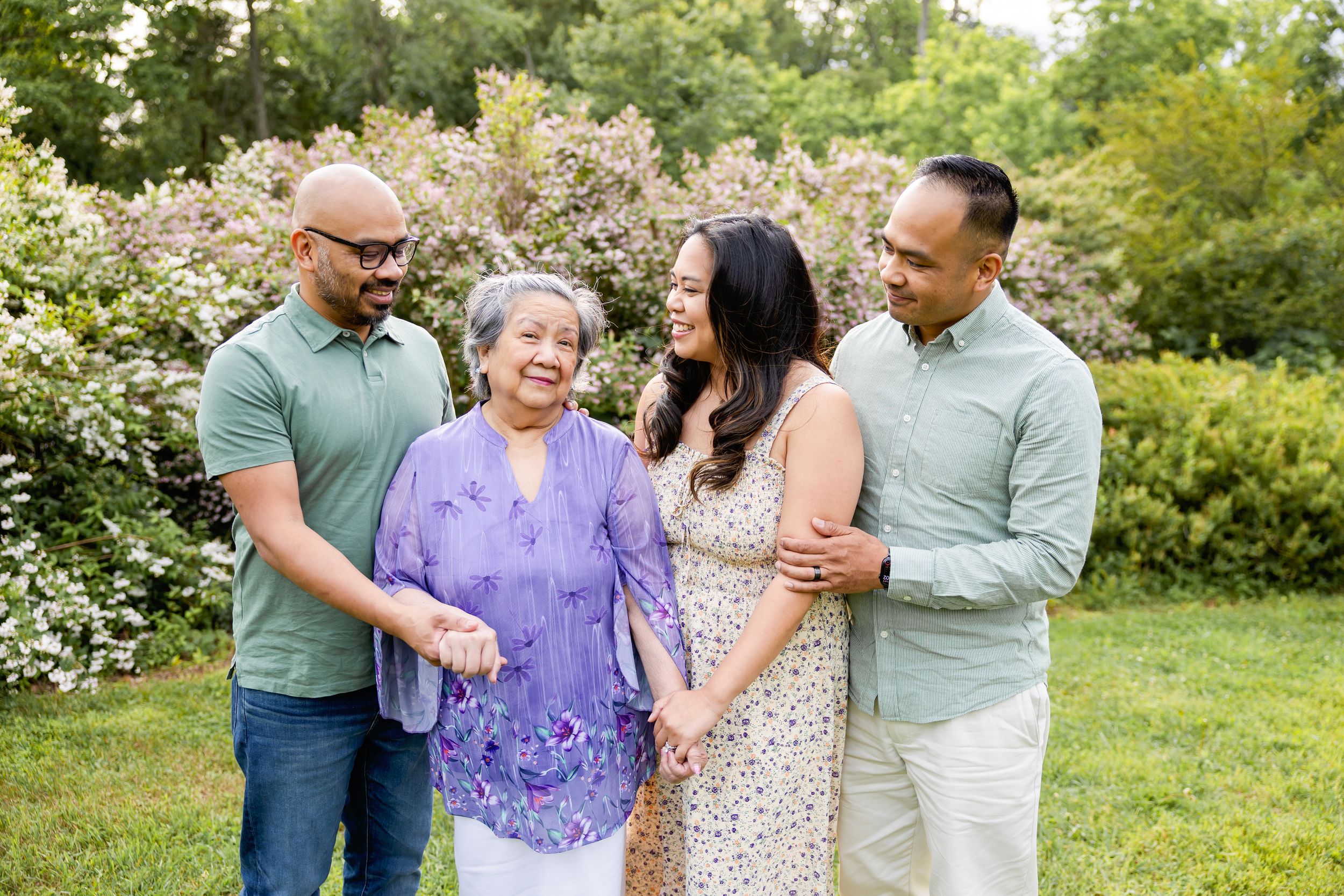 Family gathers together outdoors enjoying a sunny day in a garden with blooming flowers in the background. Family NJ photographer