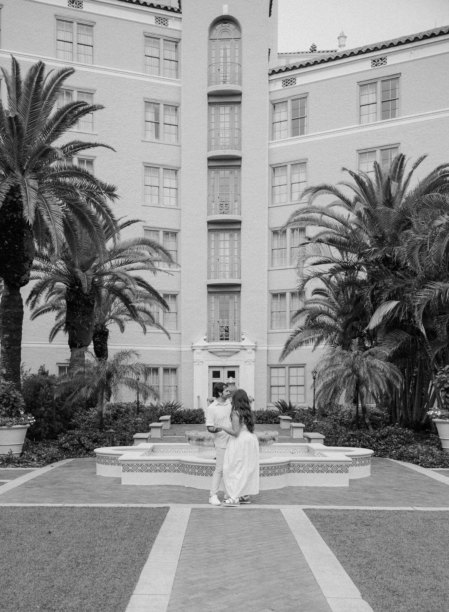 A black and white photo of a stately building courtyard with palm trees flanking a central walkway and fountain.