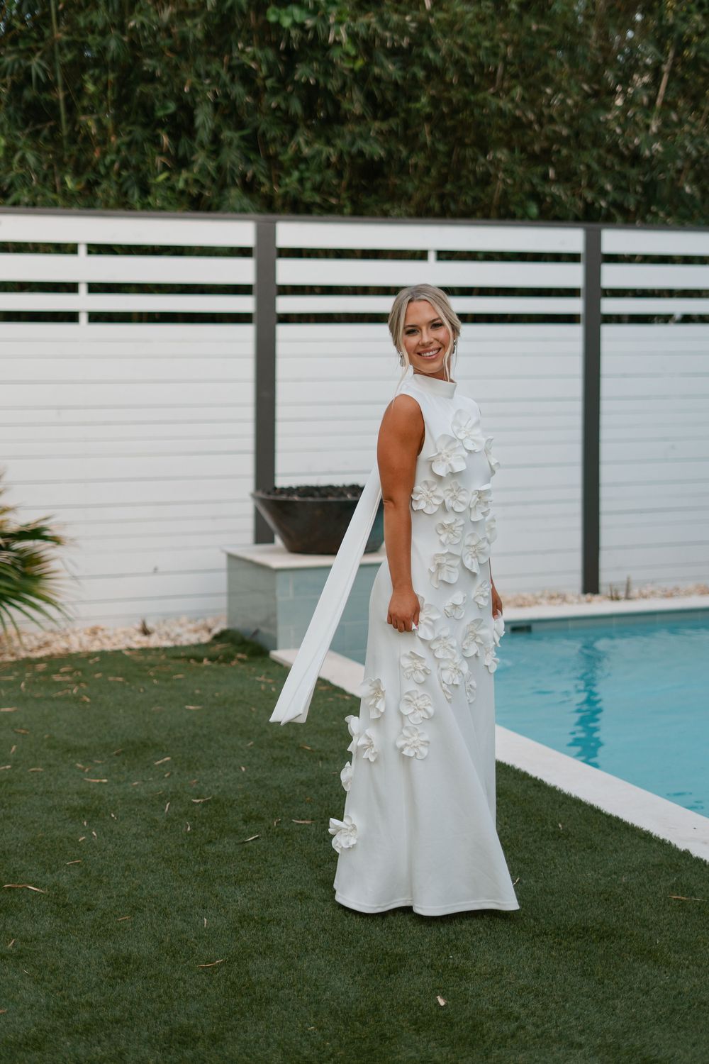 Beautiful white wedding gown with floral appliques and sleeveless design shown against backyard pool setting.