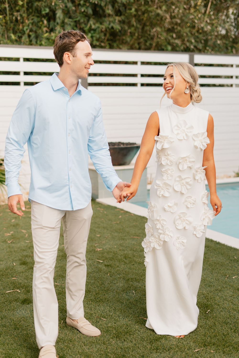 Couple walking hand in hand outdoors, with a white dress and blue shirt against a pool and white fence background.