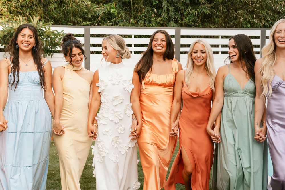 A group stands together wearing coordinated pastel and earth-toned dresses in various styles against a white fence backdrop.