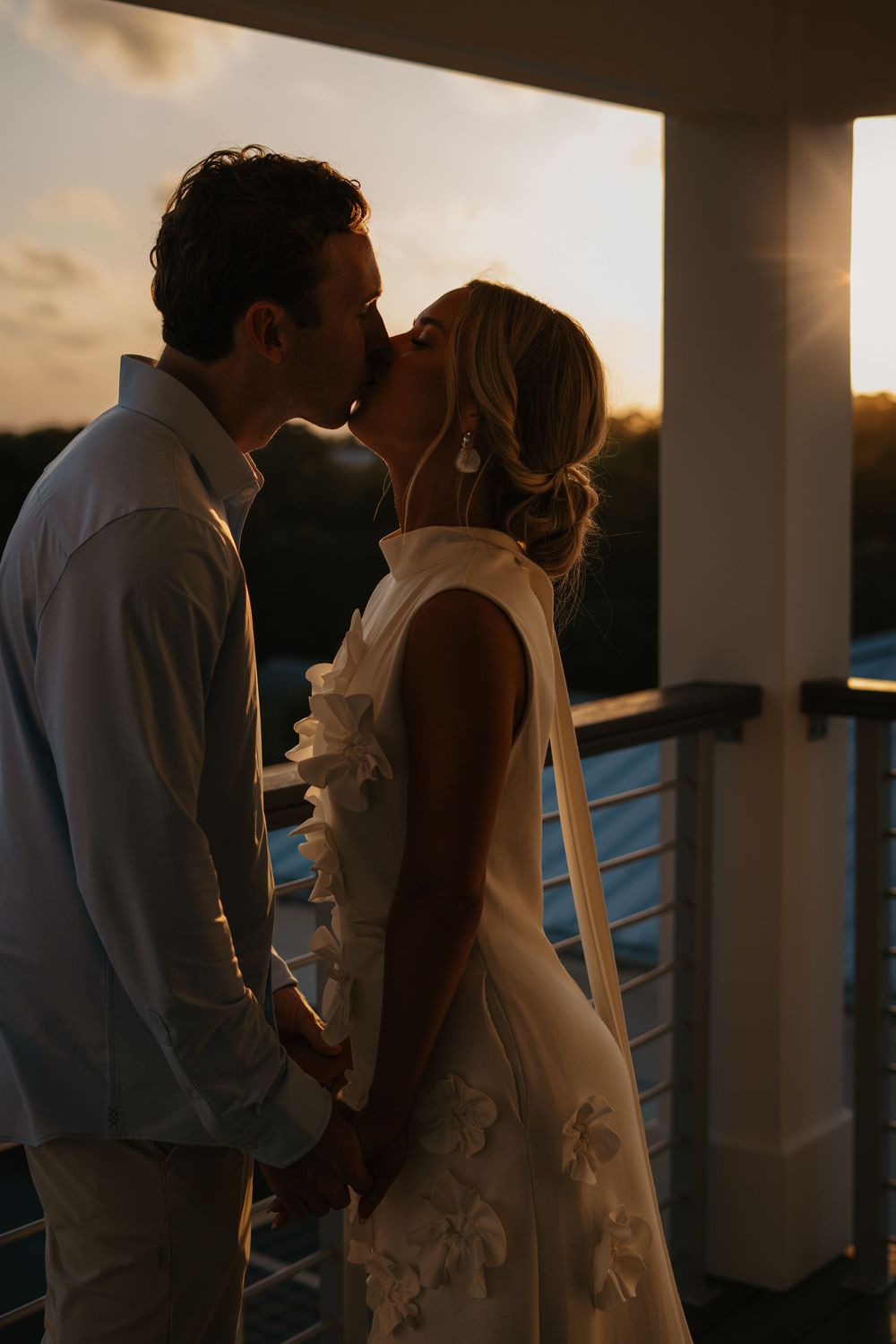A romantic sunset silhouette of newlyweds sharing a kiss on a balcony overlooking the horizon.