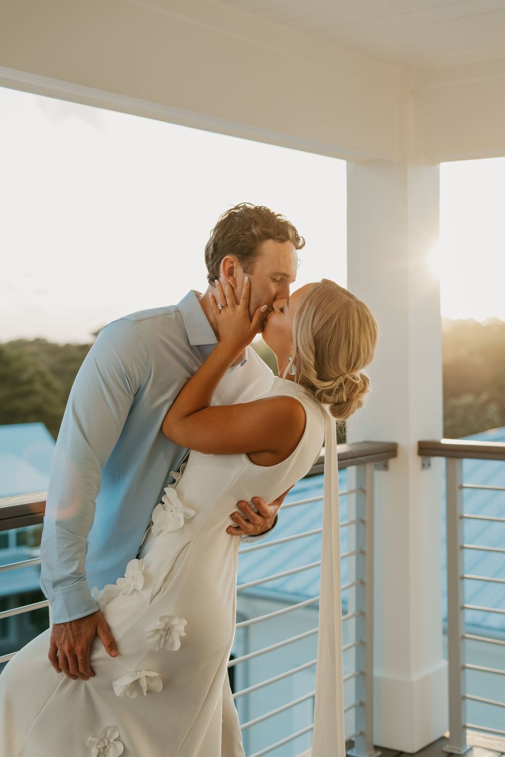 A romantic couple shares a tender moment at sunset on a waterfront balcony, the bride wearing an elegant white dress.
