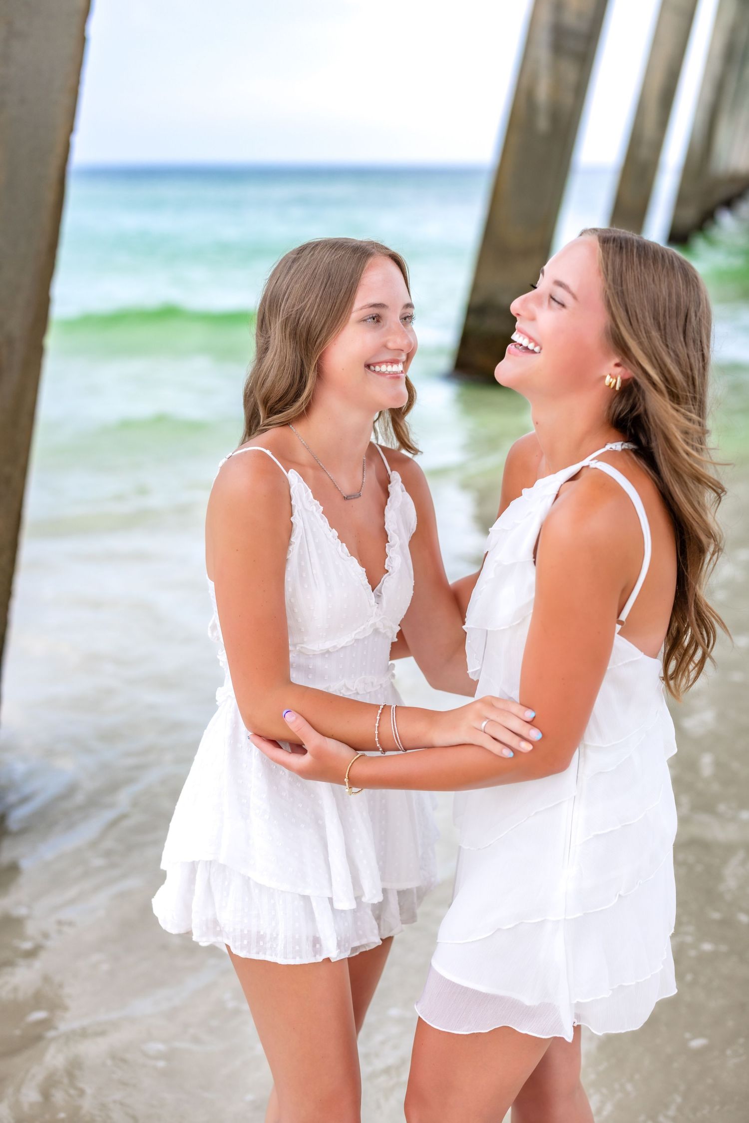 Two people in white dresses sharing a joyful moment together while standing on a beach near ocean waves.