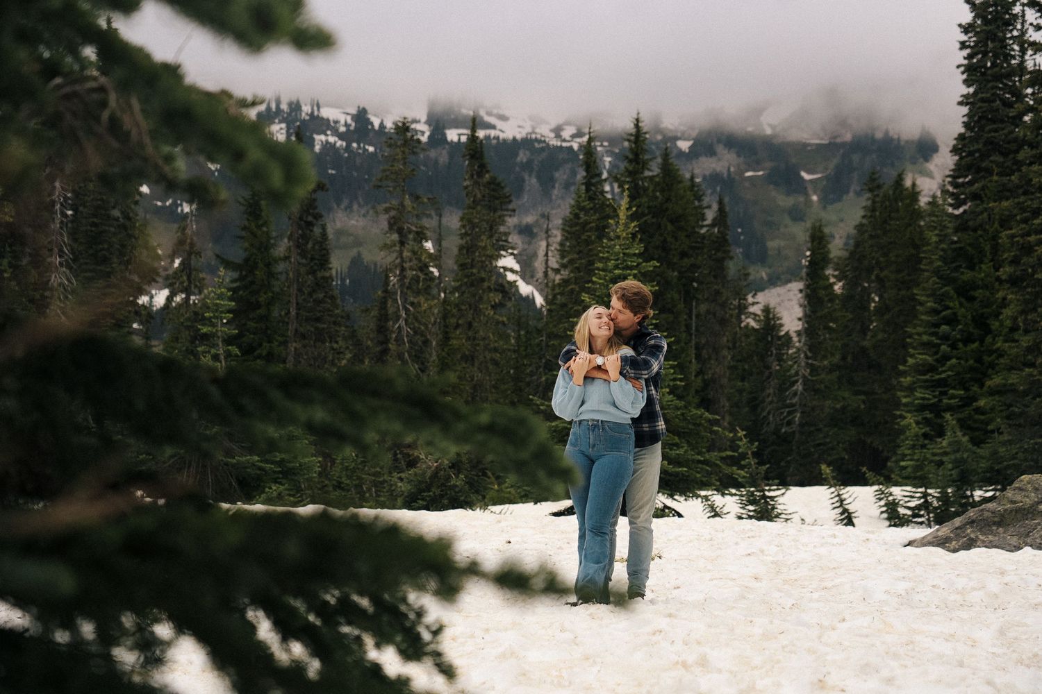 Couple embraces while standing in snowy mountain landscape surrounded by evergreen forest and misty peaks.