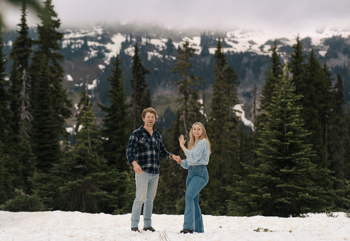A couple stands together in the snow surrounded by evergreen trees and mountains in the background.