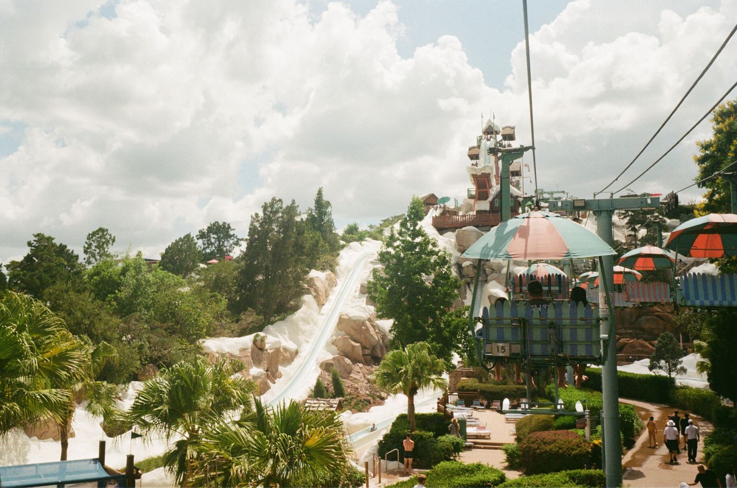 Ski lift chairs transport visitors over lush tropical terrain at a mountain resort under bright blue summer skies.