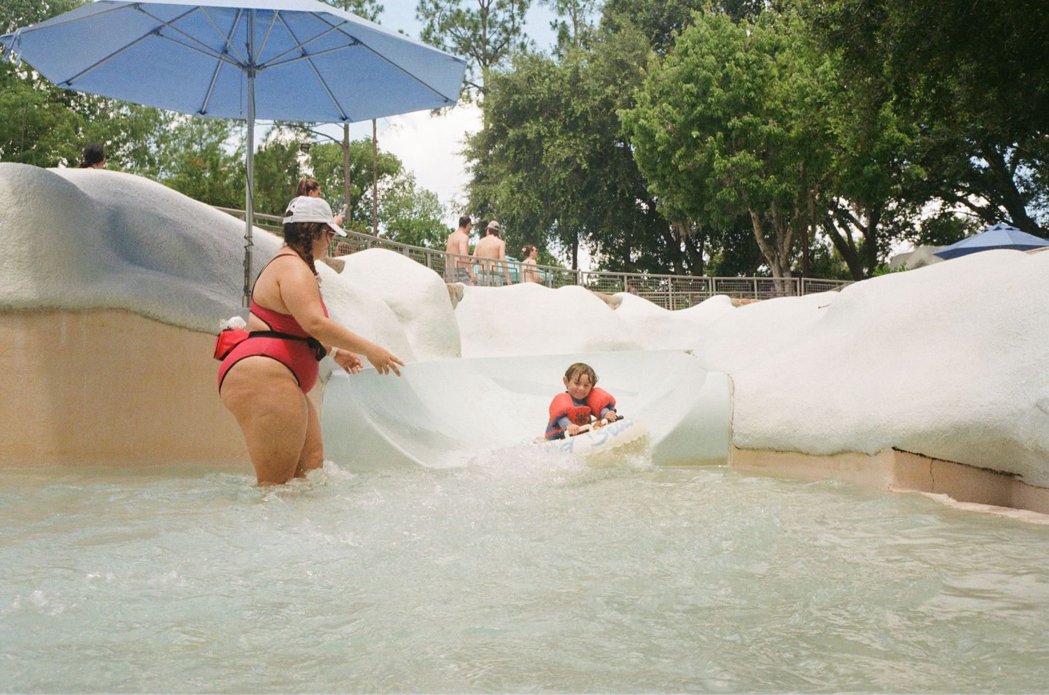 People enjoy cooling off at a water park's white curved slide feature with blue umbrella shade on a sunny day.