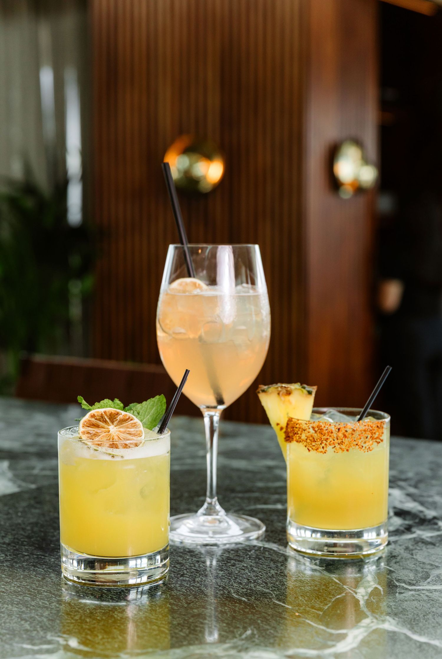 Three elegant citrus cocktails with garnishes lined up on a dark marble bar counter in warm lighting.