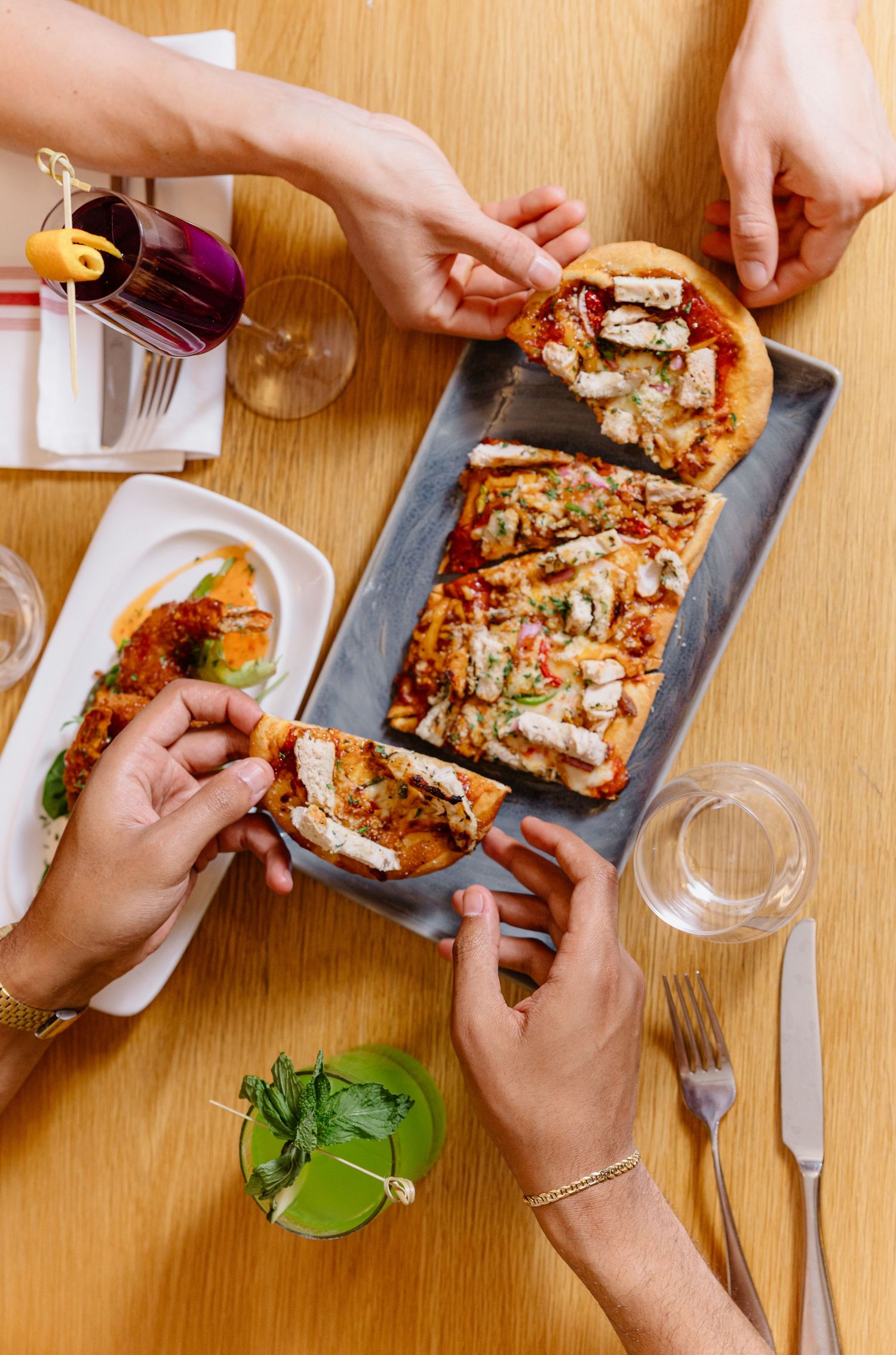 Hands reaching for slices of flatbread pizza served on a gray platter with wine glasses and herbs on wooden table.