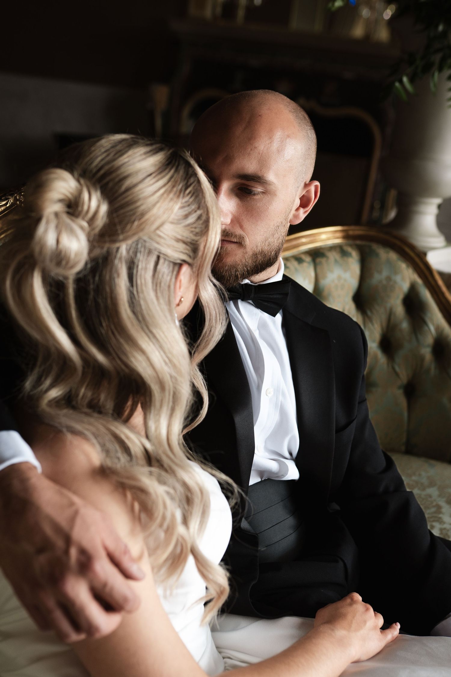 A couple in formal wedding attire pose in an elegant room with a chandelier and vintage furniture among floral arrangements.