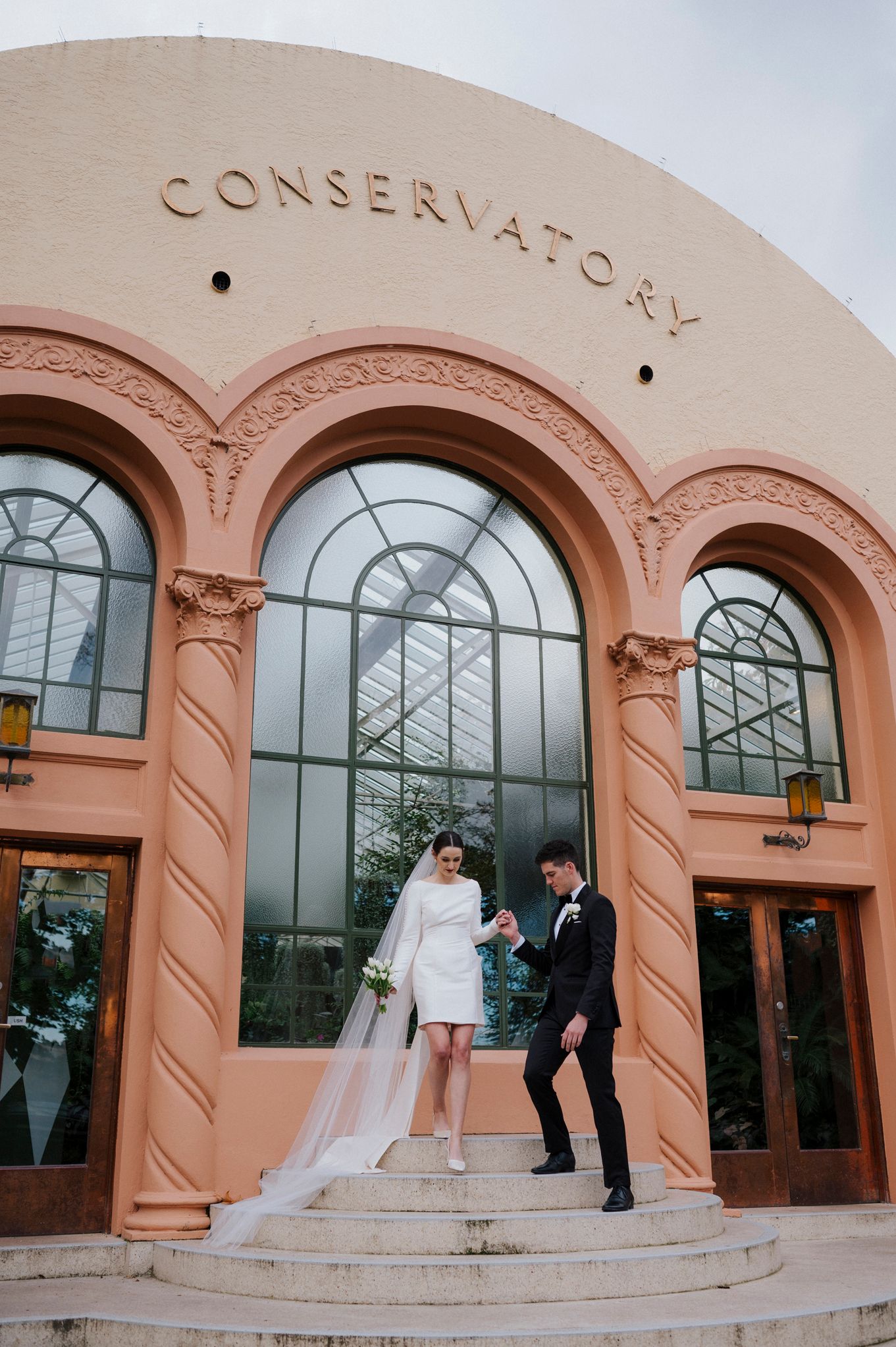 Newlyweds pose on steps outside a coral-colored conservatory building with arched windows.
