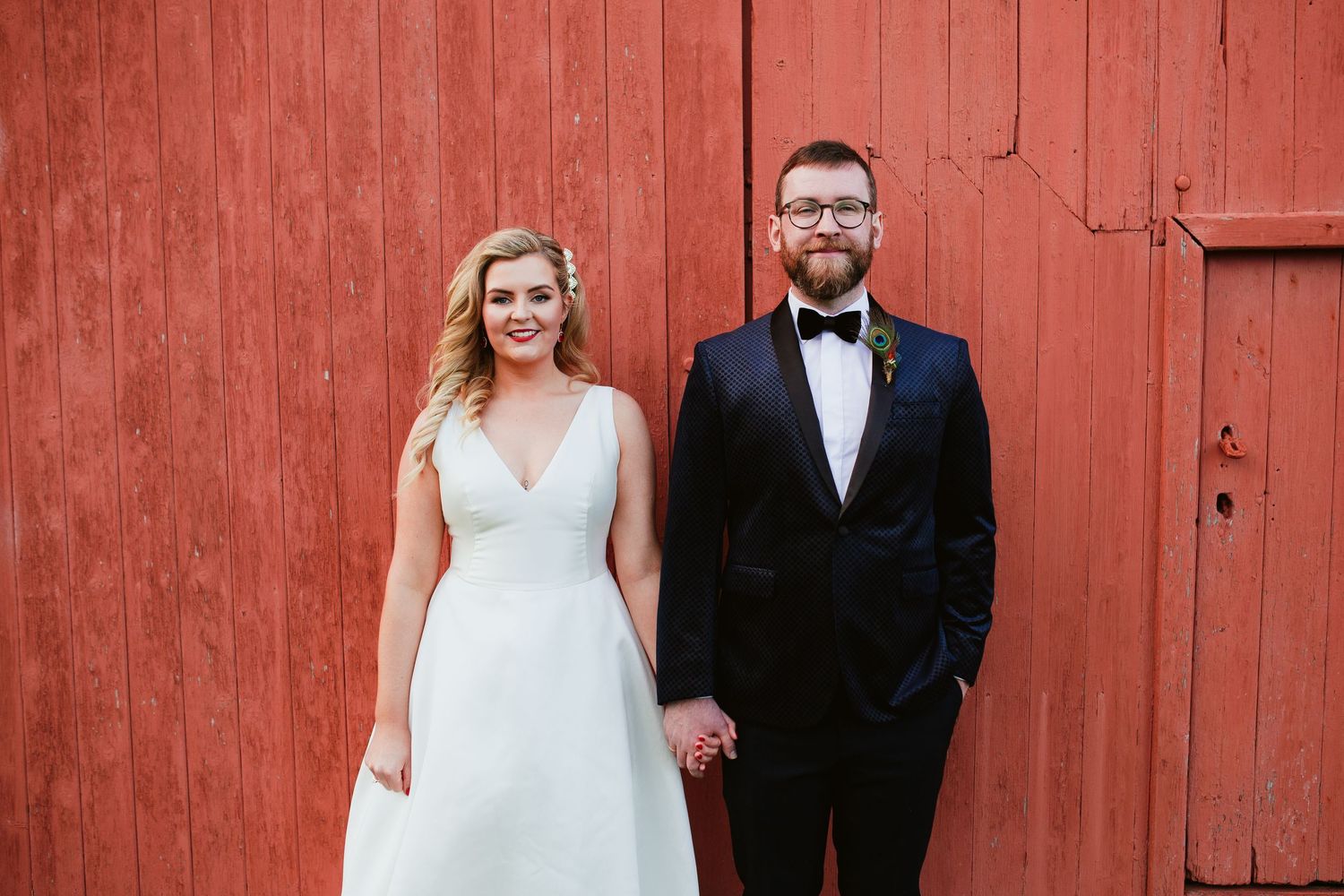 A wedding portrait of a couple standing against a red rustic barn wall.