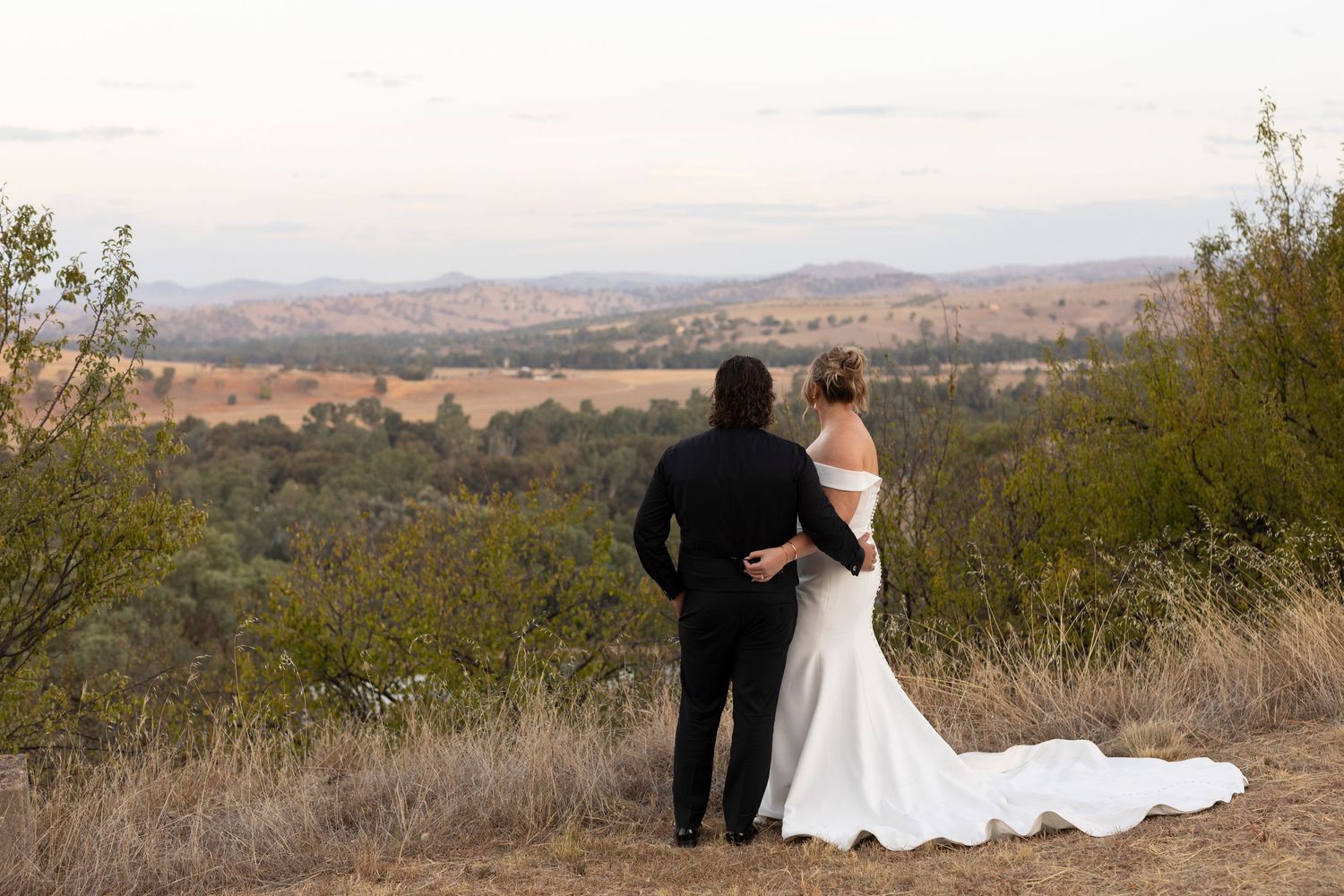 Newlywed couple stands together on hilltop overlooking scenic valley landscape at sunset.
