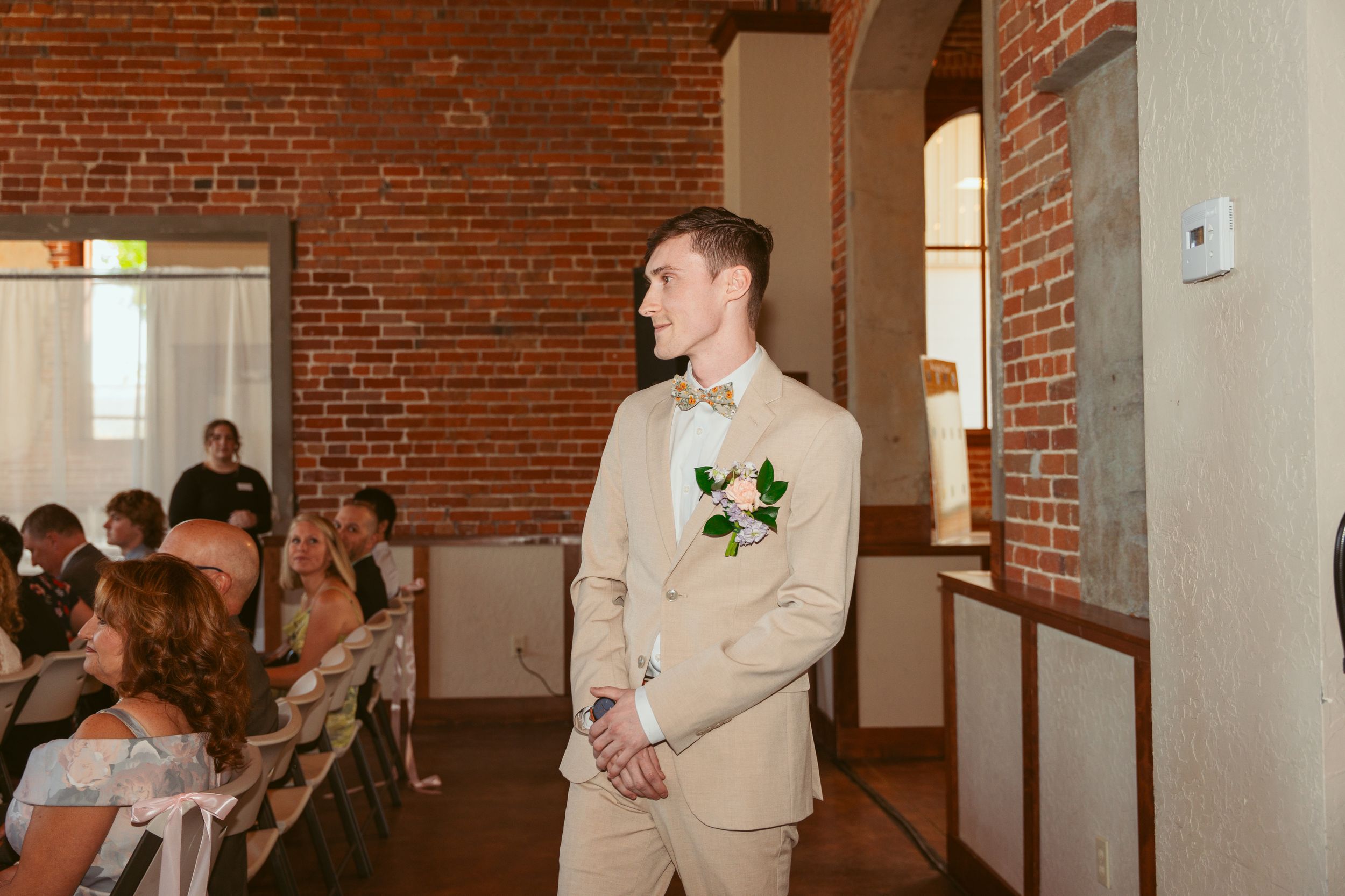 Wedding guests gather in a rustic brick venue while groomsmen in tan suits line up along the wall for a ceremony.