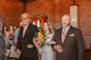 A bride in a white dress holds a colorful orange and peach floral bouquet while walking down the aisle at a rustic brick venue.