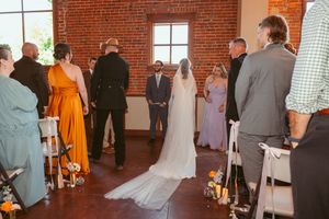 Indoor wedding ceremony in a rustic brick venue with bridesmaids in orange dresses and guests gathered around.