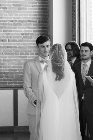 A wedding ceremony takes place in an industrial venue with exposed brick walls and natural light streaming through windows.