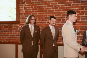Three men in formal suits stand together in a brick-walled room during a wedding ceremony.