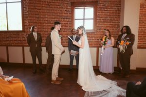 Couple exchanges vows during wedding ceremony in rustic brick-walled venue with wedding party looking on.