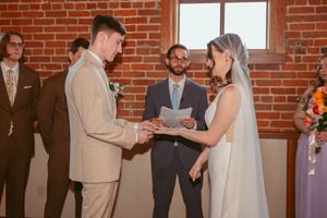 Wedding ceremony at a rustic brick venue shows hands exchanging rings during an intimate indoor celebration.