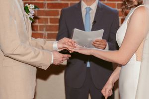 Close-up of hands exchanging wedding rings during intimate ceremony with officiant and couple in front of brick wall.
