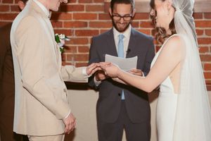 Wedding ceremony with couple exchanging rings in front of brick wall and windows.