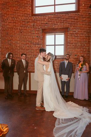 Bride and groom share their first dance on a dark wooden floor in an industrial-style brick venue with natural lighting.
