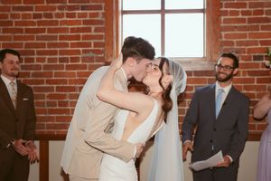 A romantic wedding couple shares their first kiss in a rustic brick-walled venue while guests look on with joy.