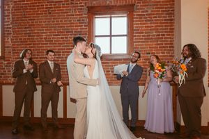 Newlyweds share first kiss during wedding ceremony in rustic brick venue with wedding party watching.