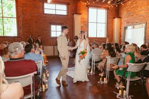 Wedding ceremony in a rustic brick venue with string lights overhead as couple walks down aisle lined with white chairs.