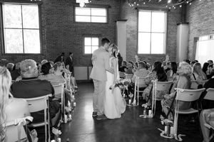 Black and white photo of an indoor wedding ceremony with guests seated in rows of white chairs inside a brick-walled venue.