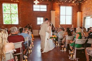 Couple shares their first kiss during wedding ceremony in industrial brick venue with natural light streaming through windows.