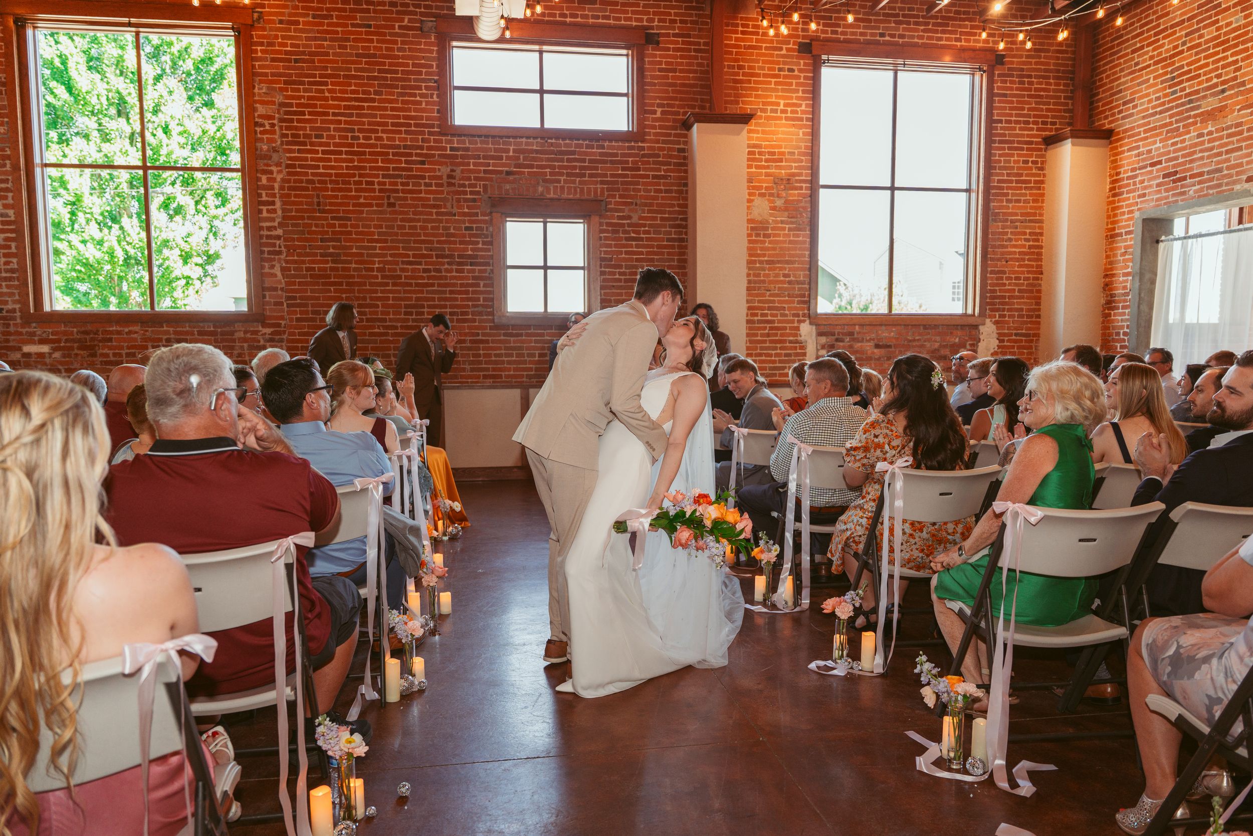 A wedding couple shares their first kiss in a rustic brick venue surrounded by seated guests.