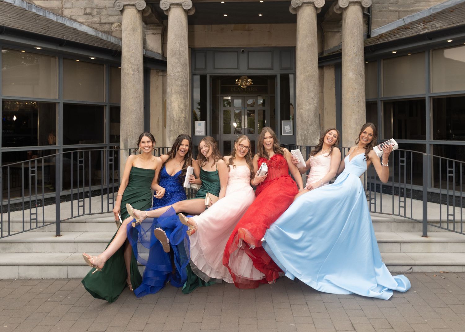 A group wearing formal gowns in different colors pose together in front of a columned building entrance.