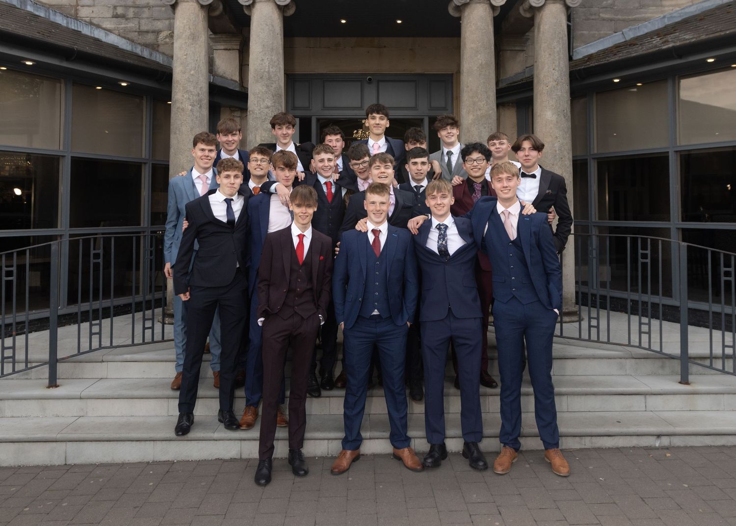 A group wearing matching dark suits and ties pose formally in front of a columned building entrance.