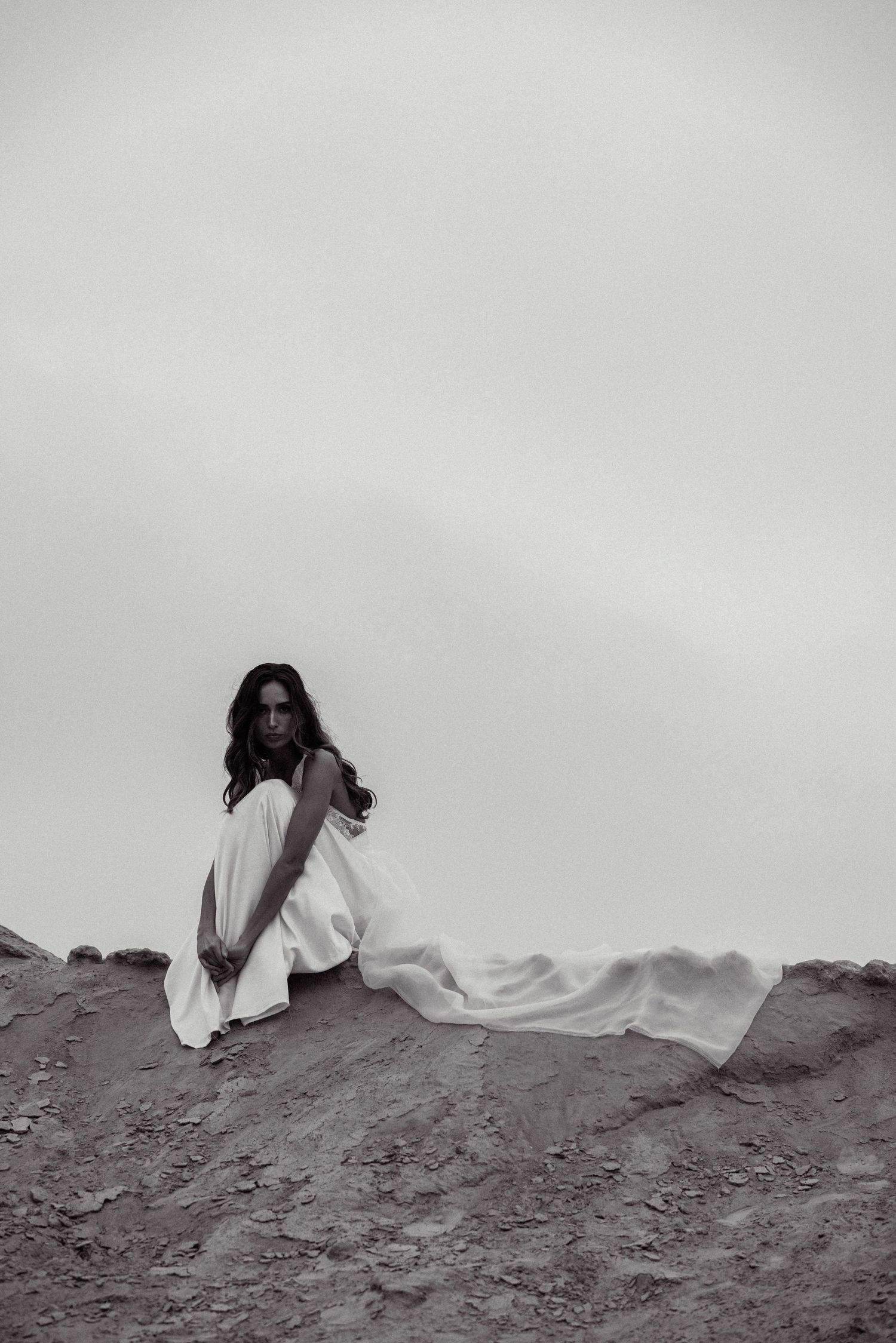 A dramatic black and white photo shows a woman in white dress sitting thoughtfully on a sandy beach.