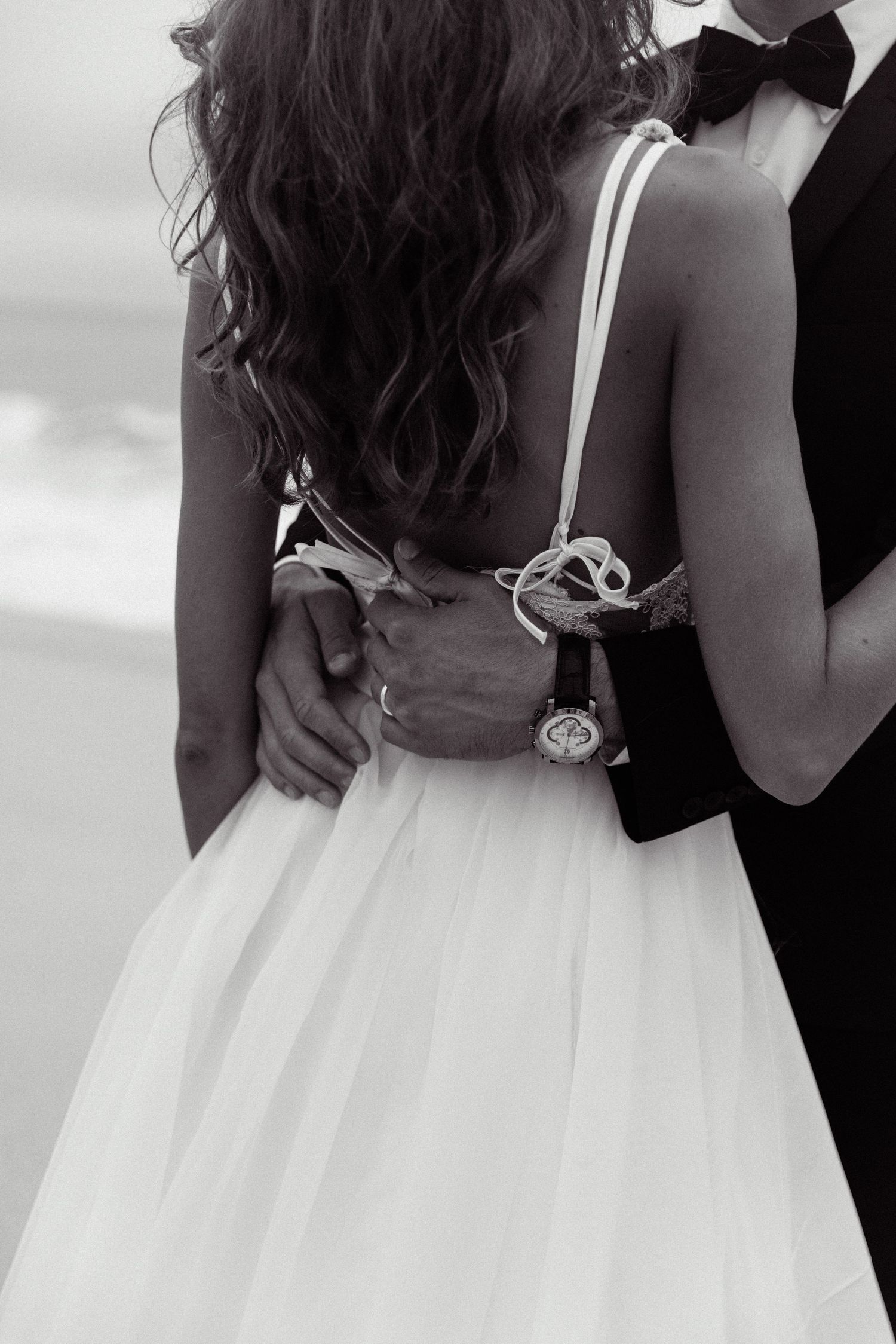 A close-up black and white photo of a couple embracing, showing the back detail of an elegant white wedding dress.